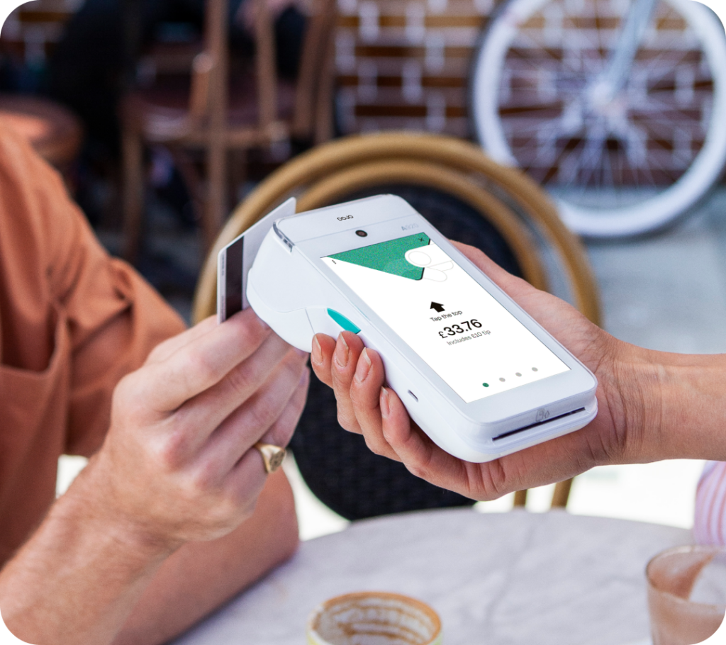 A person inserts a card into a white, handheld payment terminal displaying a transaction of £32.78 on its screen. They are seated outdoors at a café, with chairs and a table visible in the background.