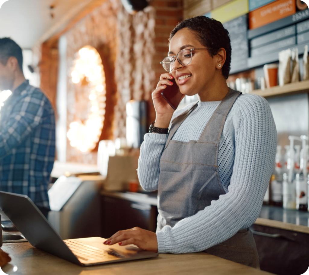 A person in a grey apron and glasses is smiling while talking on the phone in a café. They are standing at a counter, looking at a laptop. Another person is blurred in the background, wearing a checked shirt.