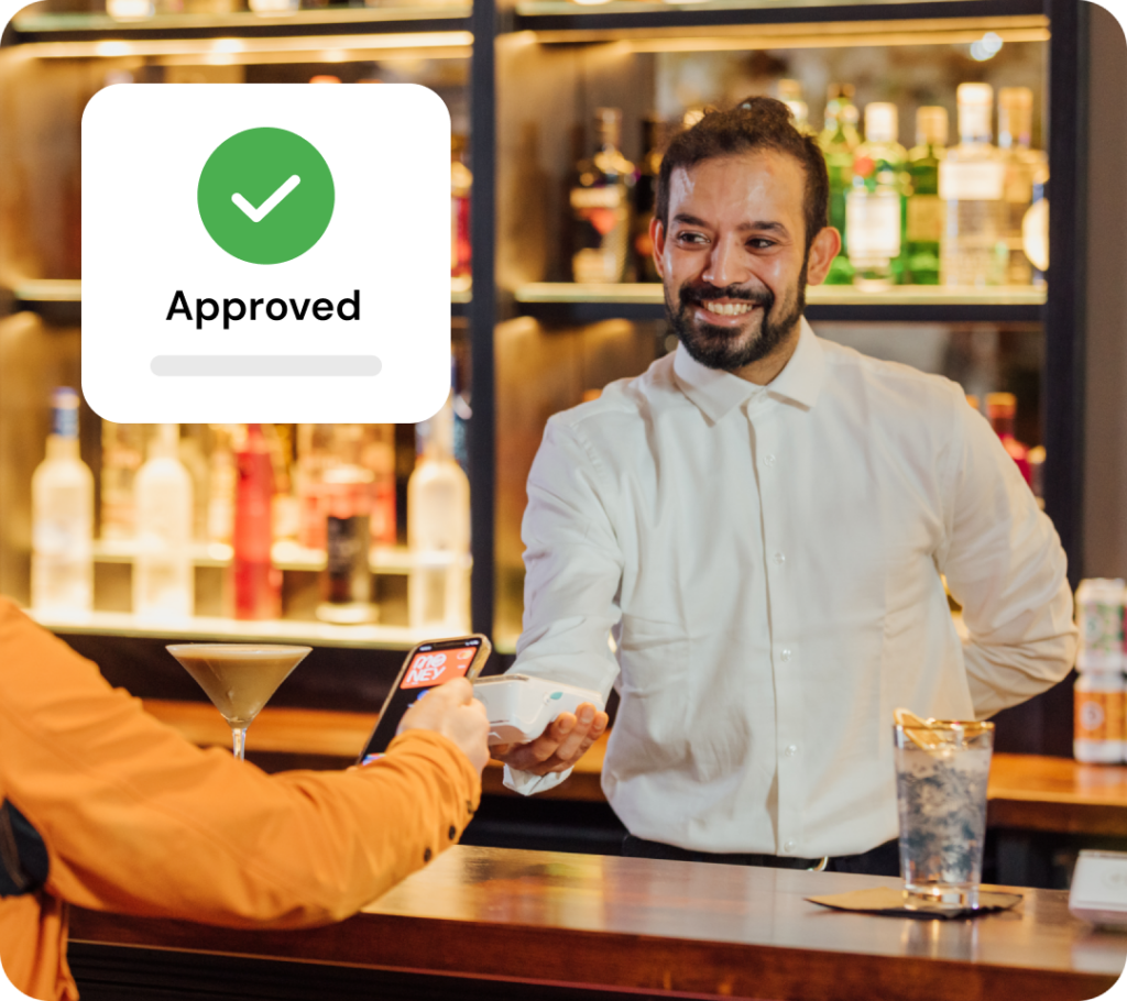 A barman in a white shirt smiles as he receives payment from a customer holding a smartphone. A drink with ice and a cocktail are on the bar. A digital Approved notification overlays the image.