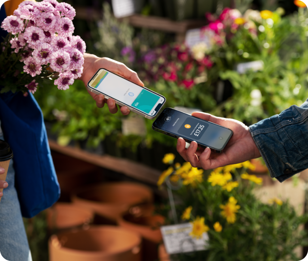 Two people exchanging a contactless payment in a garden centre. One hand holds a smartphone displaying a payment app, while the other holds a phone showing £17.35. The background features potted plants and flowers.