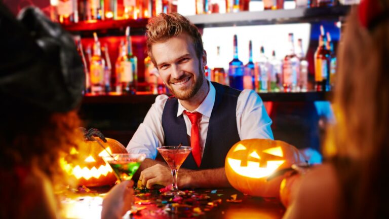 A smiling man in a waistcoat and red tie sits at an eerie bar decorated for Halloween, surrounded by lit jack-o-lanterns and colourful cocktails. The background showcases shelves of bottles, with confetti sprinkled across the counter, adding to the charm of the festive venue.
