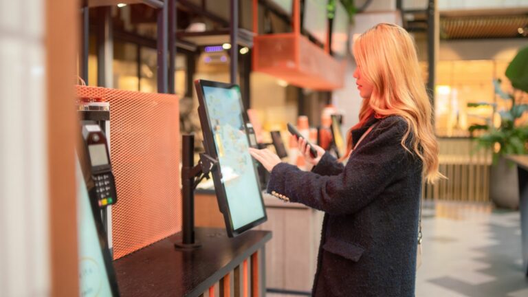 A woman with long hair interacts with a touchscreen kiosk, showcasing innovation in this modern setting. She appears to be making a business purchase or order, holding a smartphone. The background is softly blurred, accented by warm lighting and lush plants.