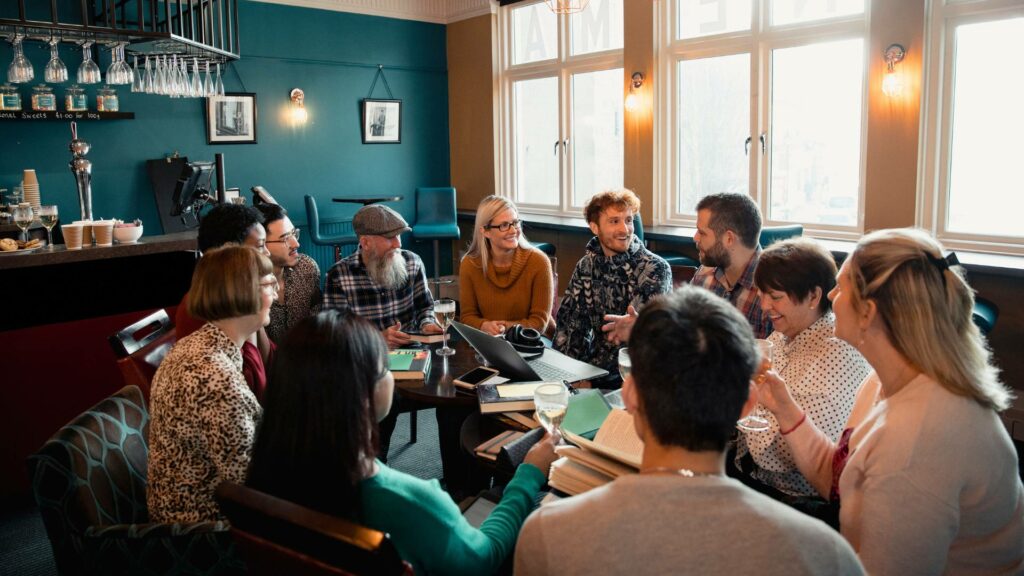 A diverse group of people sit around a table in a cosy pub, engaged in a lively discussion like members of exclusive clubs. Laptops, notebooks, and drinks are on the table. The interior features teal walls, framed art, and large windows letting in natural light.