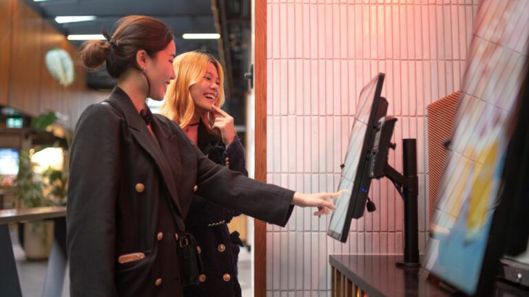 Two women stand at a self-service kiosk in a modern indoor space, embodying the essence of hospitality technology. One woman touches the screen while the other watches, both smiling, as they navigate the EPOS system. The setting features tiled walls and warm lighting.