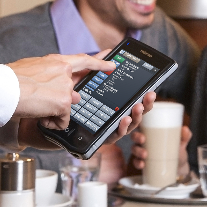 A person in the Liverpool City Region is using a handheld ordering device at a café, with the menu displayed on the screen. In the background, another patron enjoys their coffee drink surrounded by various crockery items.
