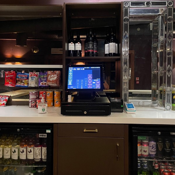 At the centre of the Liverpool City Region ambience is a bar counter with a touchscreen till. Behind it, shelves house wine bottles, while a fridge below showcases refreshing drinks. On the left side, snacks represent a diverse range of bar offerings, capturing the local flavour.