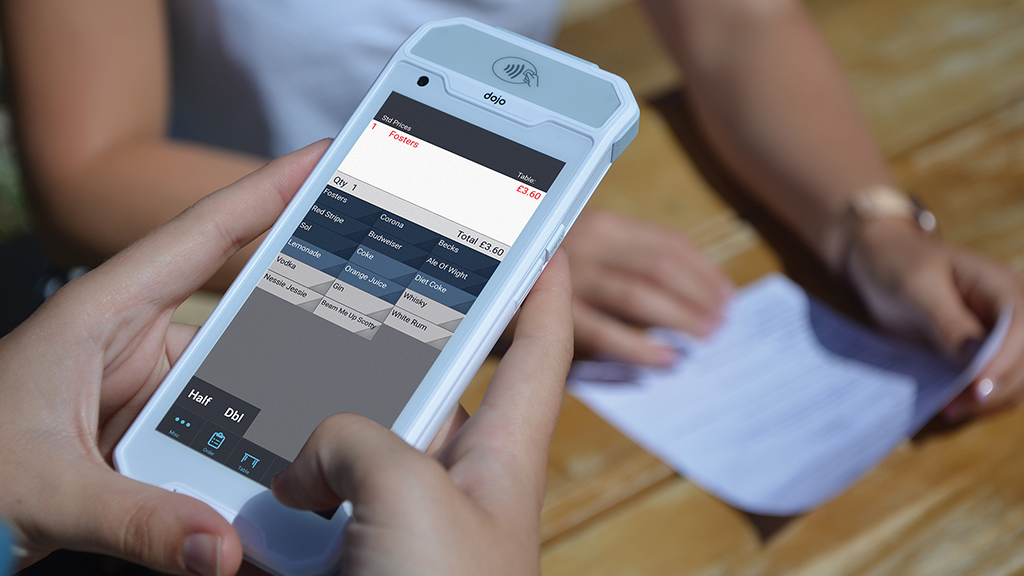 An individual holds a device displaying a menu with prices, powered by handheld EPOS software. In the background, another person prepares to receive a piece of paper on a wooden table, illustrating a seamless transaction setting.