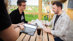 Two men sit at a wooden outdoor table in the heart of the Liverpool City Region, browsing menus. Nearby, a person holding a PocketTouch tablet cheerfully takes their order. A drink with ice rests on the table. The open patio area, surrounded by grass and picnic tables, creates an inviting atmosphere.