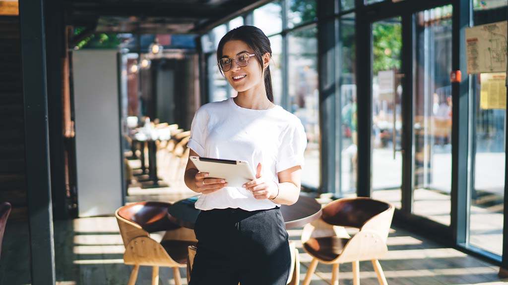 A woman in a white shirt and glasses stands inside a bright café in the Liverpool City Region, holding a PocketTouch tablet. She is smiling and looking away from the camera. The café features wooden tables and chairs, with large windows letting in natural light.