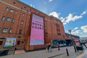 Street view of Liverpools Royal Court Theatre, showcasing its red-brick façade and a large vertical sign. People stroll by on a sunny day with scattered clouds overhead, as posters adorn the left side of this iconic building.