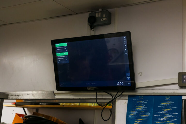 A wall-mounted digital screen displaying a user interface with various options and a clock showing 12:56. Below the screen is a cable and what appears to be a menu or list, partially visible. The screen is in a kitchen setting.