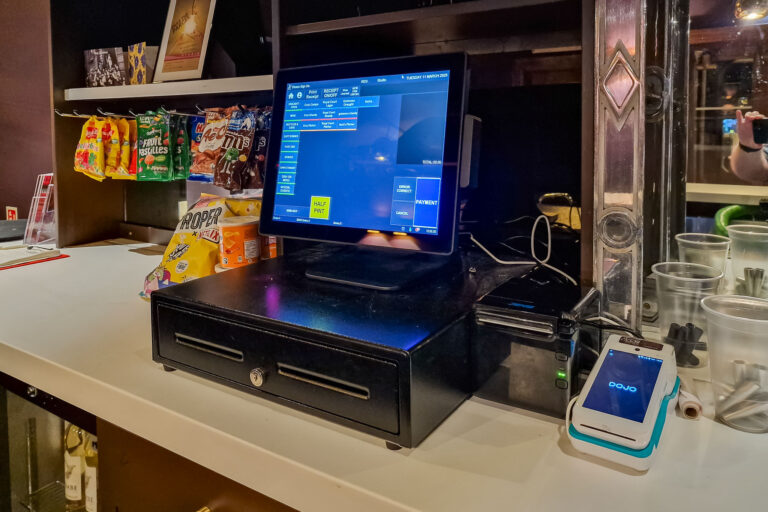 A retail counter with a touchscreen point-of-sale system on a cash drawer. Nearby are snacks like crisps and sweets. A smartphone with a payment app is on the side, and plastic cups with straws are visible in the background.