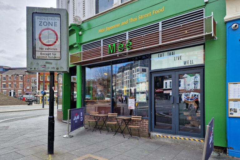 A green and brown shopfront for Meé Japanese & Thai Street Food, featuring a glass window displaying Eat Thai Live Well. Outside, a small table with chairs beckons you to savour spicy new dishes in this pedestrian area.