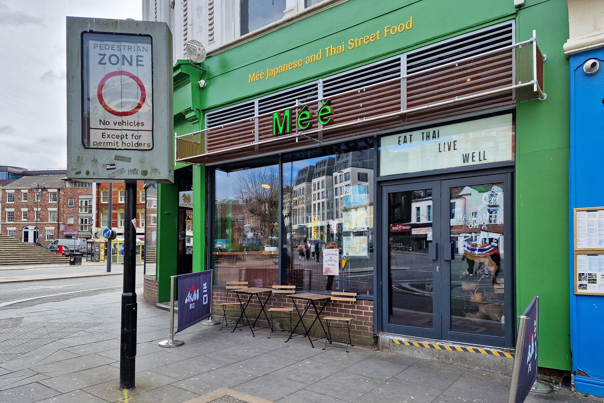 A green and brown shopfront for Meé Japanese & Thai Street Food, featuring a glass window displaying Eat Thai Live Well. Outside, a small table with chairs beckons you to savour spicy new dishes in this pedestrian area.