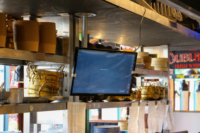 A restaurant kitchen with stacks of bowls and dishes on metal shelves. A touchscreen monitor is mounted above the shelves, displaying a digital screen. Order tickets are clipped below the screen. Reflections can be seen on the glass.