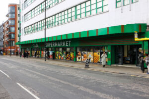 Street view of Taza Supermarket with a green and white exterior. The shopfront displays vibrant images of produce. People stroll past, enjoying a smooth shopping experience thanks to the integrated EPOS that reduces queues efficiently. A modern block of flats is on the left.