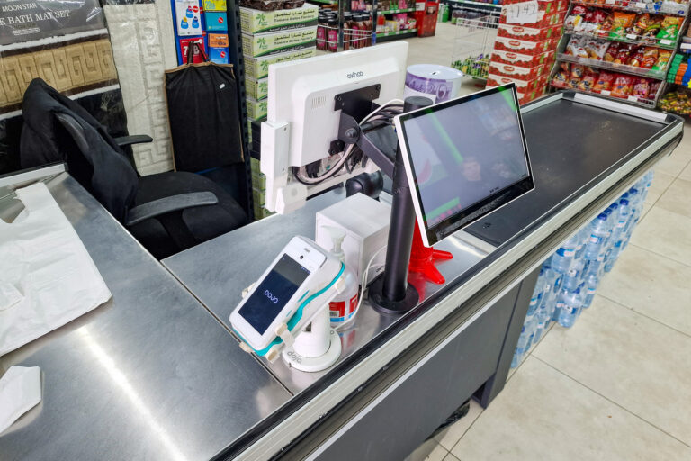 A supermarket checkout counter featuring a till, debit card machine, and a customer display screen. Nearby is a stack of paper bags, a swivel chair, and bottled water on the floor. Shelves stocked with snacks are visible in the background.