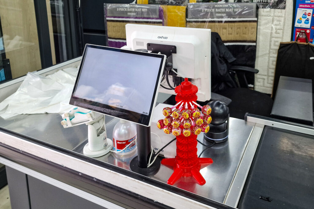 Checkout counter with a monitor, card machine, and a bottle of hand sanitiser. Next to these is a red stand holding colourful lollipops. The background shows folded bath mats on display.