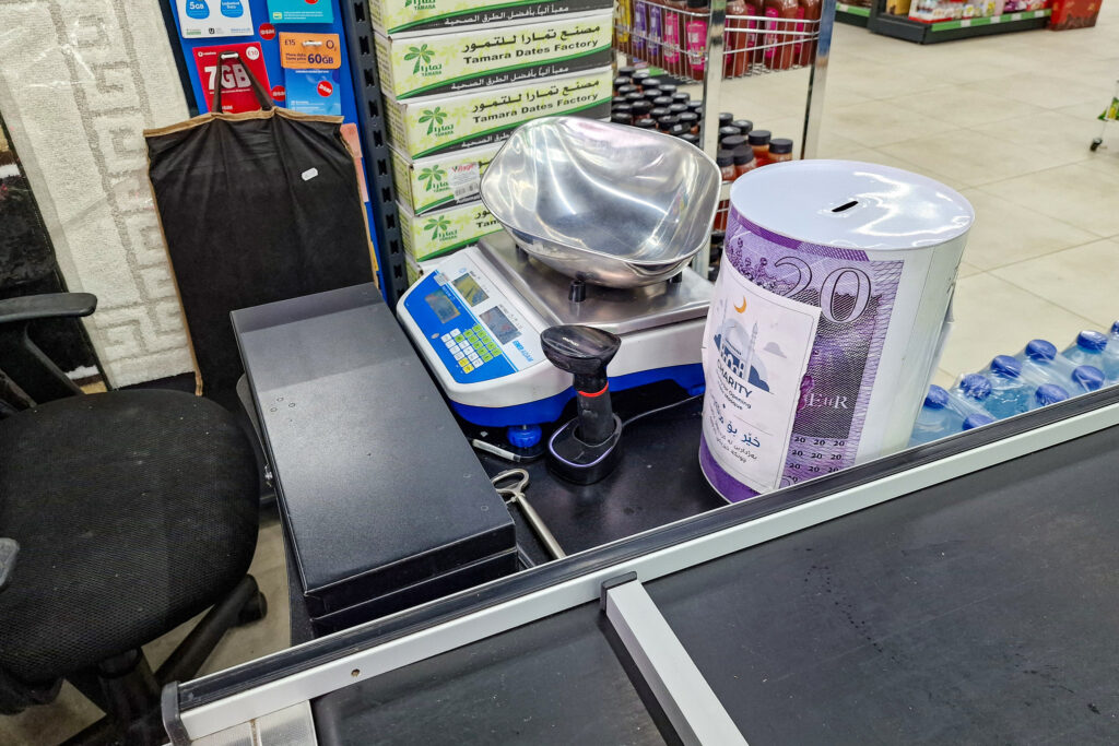 Grocery checkout counter with a digital scale and receipt printer. A donation tin featuring Arabic script is situated next to bottles of water. Shelves containing boxes of date packaging can be seen in the background.