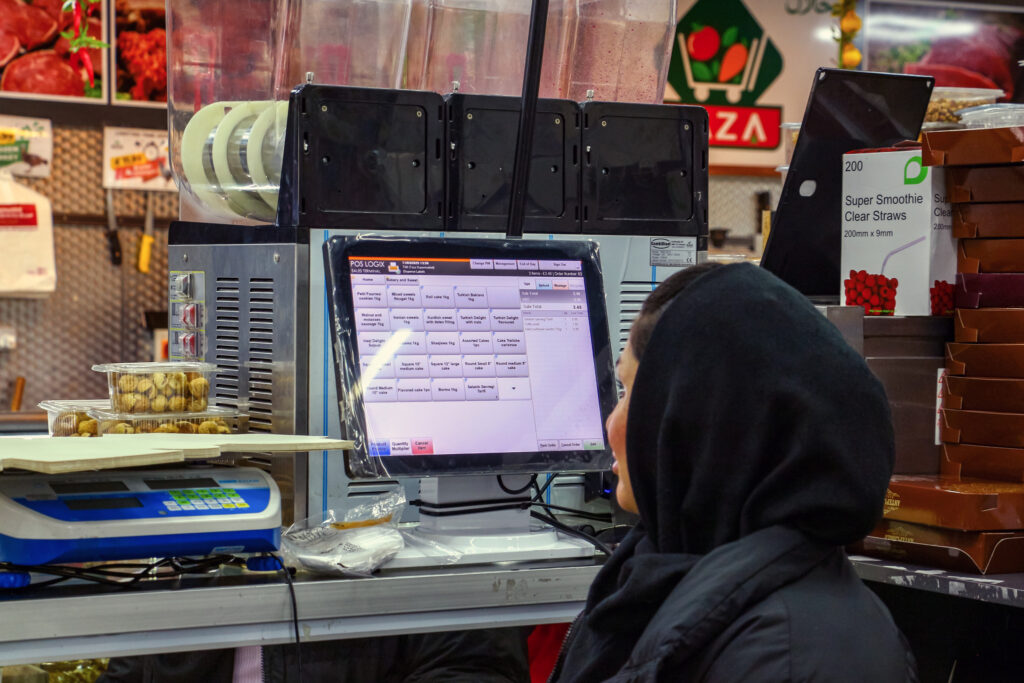 A person in a black hoodie is looking at a digital order screen at a food counter. Behind the screen, there are stacks of packaged goods and a visible display of raw meat in the background.
