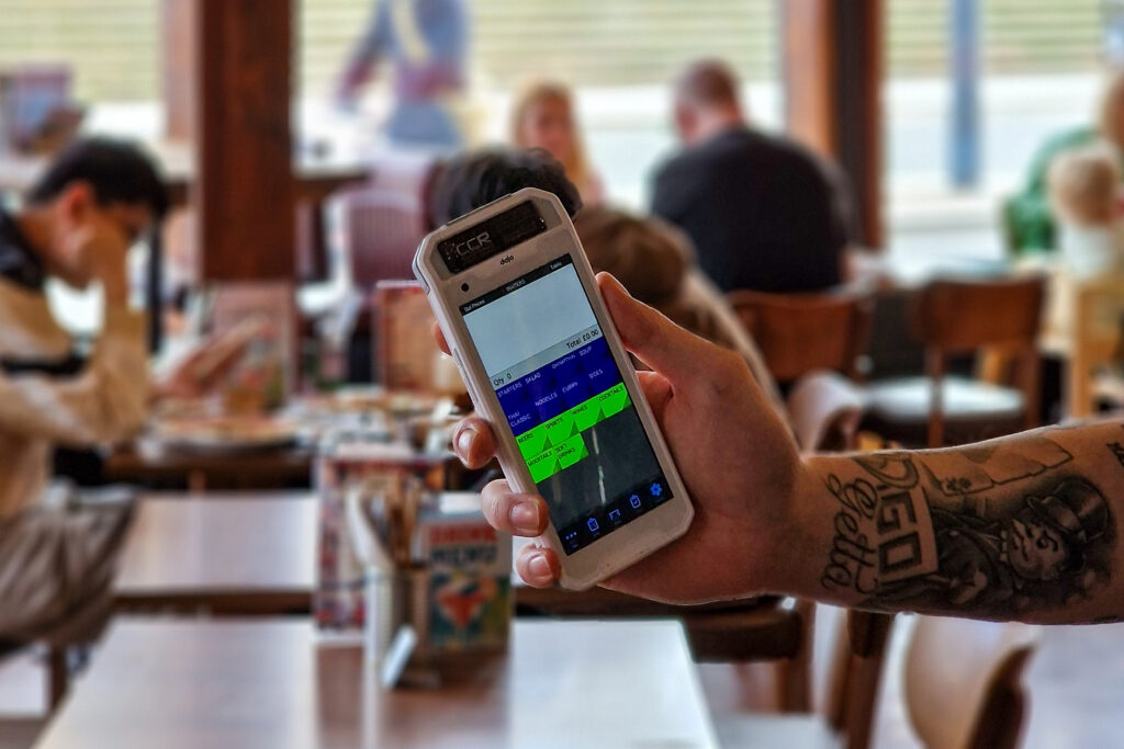 A person with tattooed arms holds a touchscreen device displaying a timetable in the bustling Liverpool City Region, where people are seated at tables in a vibrant café or restaurant environment.