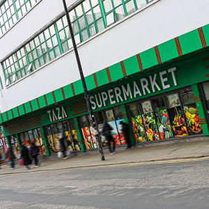 A supermarket with a green façade, named TAZA SUPERMARKET, stands proudly in the lively Liverpool City Region. Pedestrians stroll past large windows adorned with vibrant fruit and vegetable images.