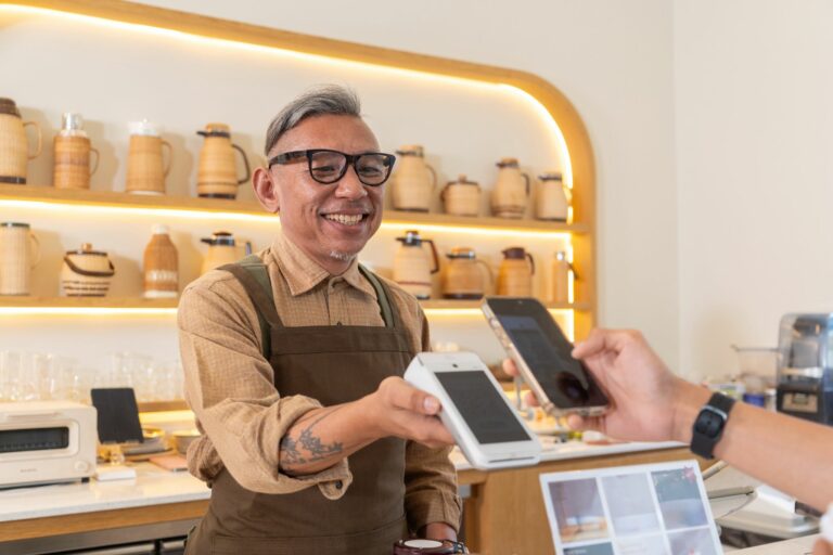 A barista in an apron hands over a Dojo Go mobile card payment device at a café with wooden shelves displaying teapots and utensils.