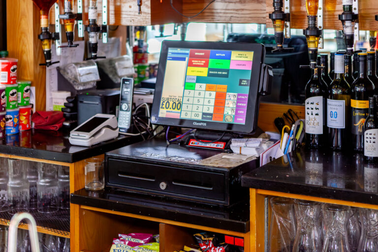 A bar counter featuring an EPOS touchscreen till, telephone, card machine, and a drawer. Bottles of wine, spirits, glasses, and snacks are displayed on shelves and counters surrounding the till.