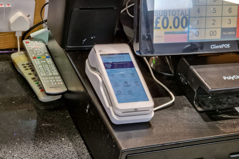 A close-up of a shop counter features a payment terminal, two remote controls, a point-of-sale till screen displaying “SUBTOTAL £0.00,” and several electronic devices on a black speckled surface.
