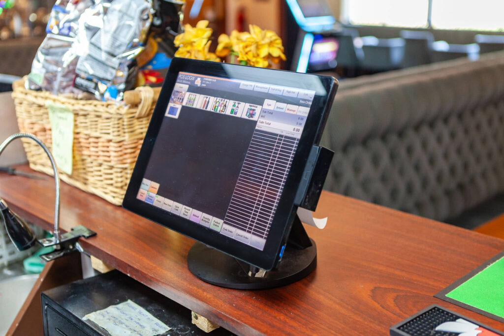A touchscreen point-of-sale system on a wooden counter, displaying a sales interface. A wicker basket with snacks and a sofa are visible in the background.