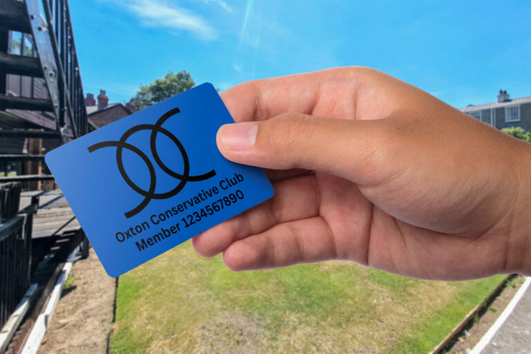 A hand holding a blue Oxton Conservative Club membership card with large, overlapping black letters OC and the text Member 1234567890, in an outdoor setting with grass, buildings, and a clear sky.