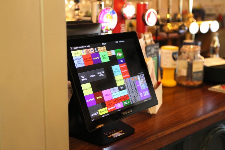 A touchscreen point-of-sale system rests on a wooden counter in a bar or restaurant, displaying various colourful menu options. Drink taps and condiment containers can be seen in the background.