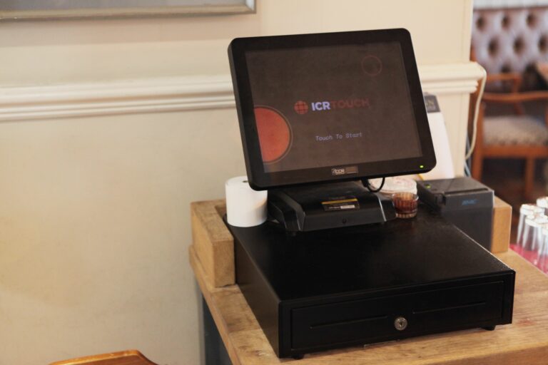A touchscreen point-of-sale system rests on a black cash drawer, with a small receipt printer and a roll of till roll beside it in a restaurant setting.