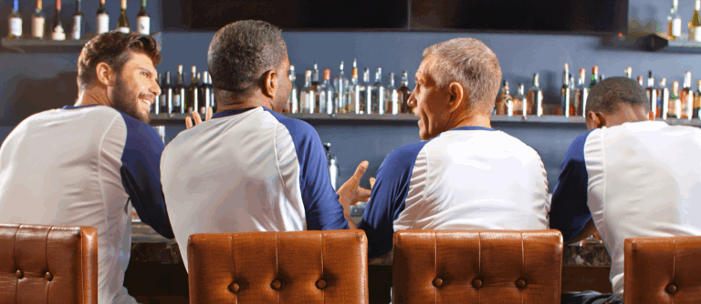 Four men in matching white and blue shirts sit on bar stools at a bar, chatting about flow management in social clubs, with shelves of bottles behind them. The men are viewed from behind.