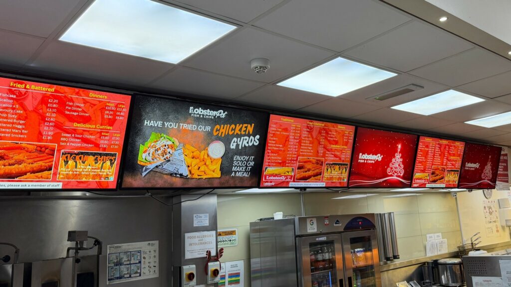 A row of digital signage menu boards above a fast-food counter displays fried foods, drinks, and special offers, with bright red and yellow accents and images of meals such as chicken gyros and fried fish.