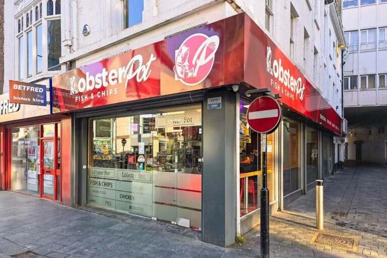 Lobster Pot, a fish and chip shop in Liverpool, has a red sign and logo, large windows, digital menus inside, and a no entry road sign beside the entrance.