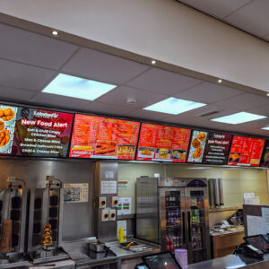 A fast-food counter with digital menu boards showing a selection of fried chicken meals, snacks, and drinks. Cooking equipment and food warmers are visible behind the counter. The restaurant looks clean and brightly lit.
