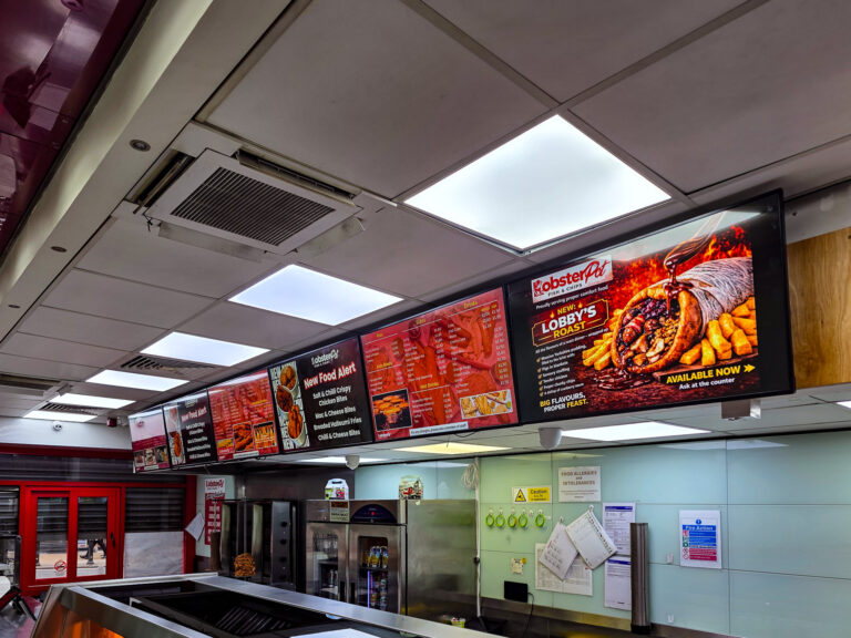 A fast food restaurant counter with illuminated menu boards showing food items, including fish, chips, and wraps. A bright sign advertises “Lobster’s Logist.” The counter and kitchen area are visible beneath the menus.