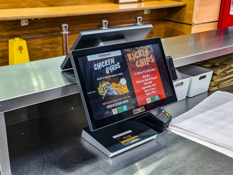 A touchscreen ordering kiosk on a restaurant counter shows menu items Chicken Gyros and Kickin Chips with various flavour options. Nearby are a card reader, paper sheets, and neatly arranged serviettes.