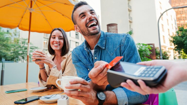 A smiling man in a denim shirt makes a contactless payment at an outdoor café, holding his cup next to a smiling woman. A bright umbrella and city buildings feature in the background, emphasising the UK’s £100 limit convenience.