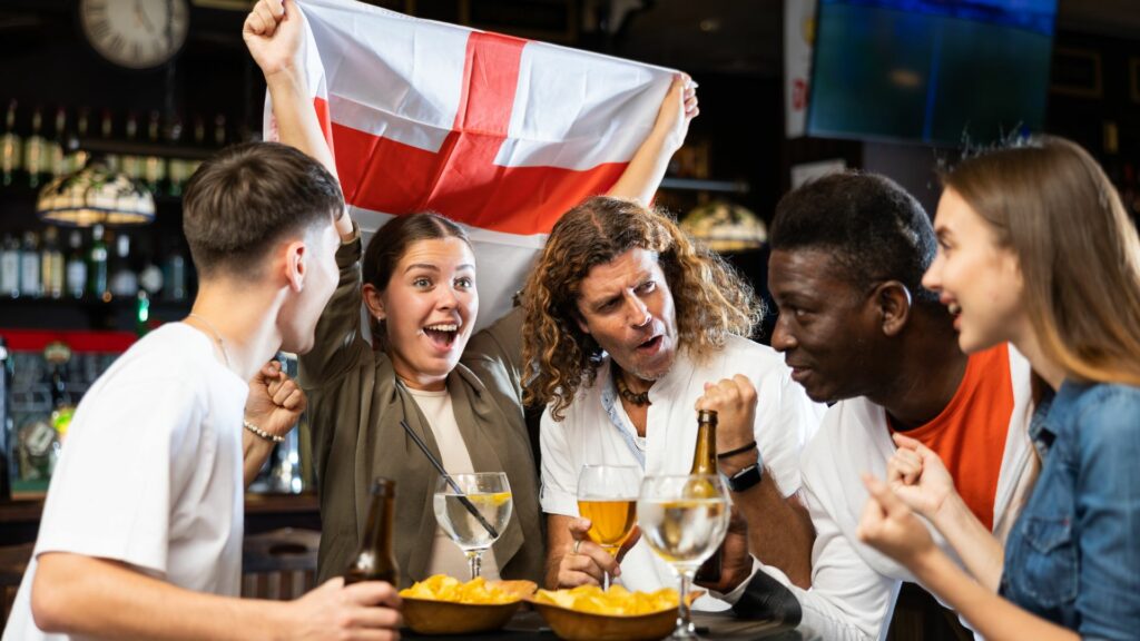A group of mates celebrate in a pub with drinks and nibbles, one waving an England flag. The buzz is tangible as they cheer on their team during the crucial stages of World Cup 2026.