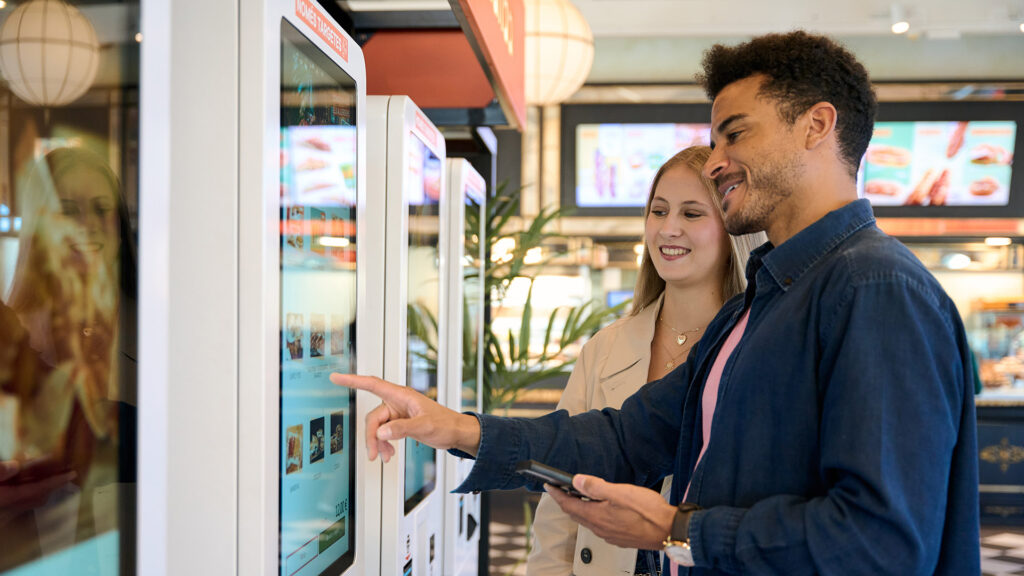 A man and woman smiling as they use a self-service kiosk in a fast food restaurant, with menu items shown on digital screens in the background.