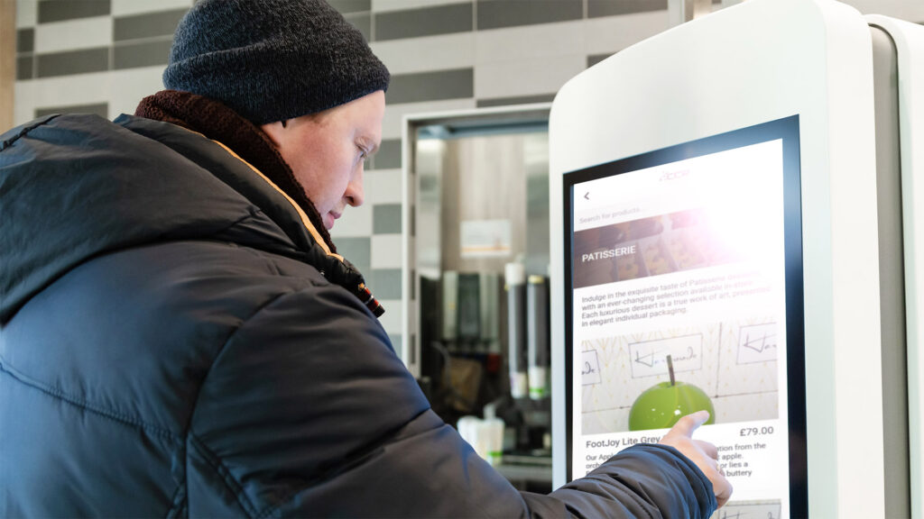 A man near self service restaurant, kiosk, terminal with empty screen.