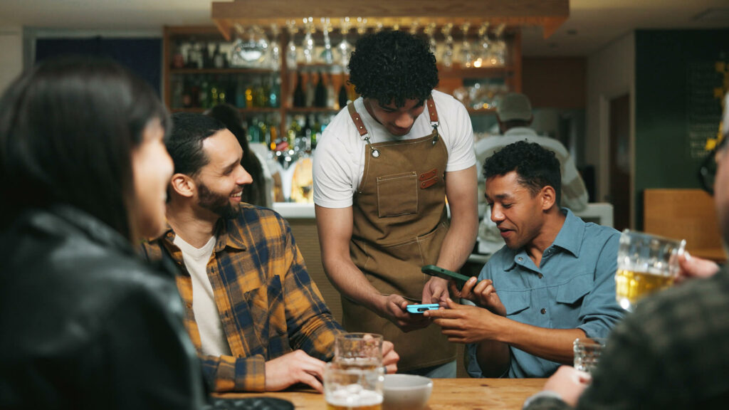 Waiter, man and phone with payment at pub for beer purchase, paperless transaction or drinking. Friends, people or server with POS machine in bar for easy banking, drinks checkout or social gathering