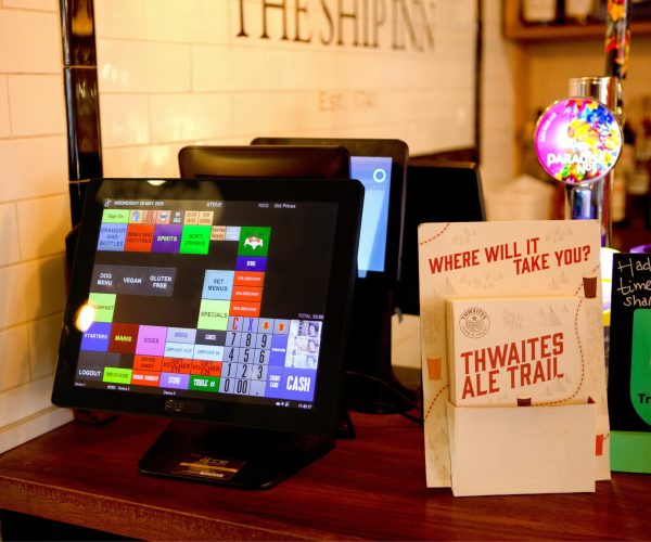 A touchscreen till sits on a bar counter beside beer pumps, a Thwaites Ale Trail leaflet, and a TripAdvisor review sign. Behind are part of a white tiled wall and bottles of alcohol.