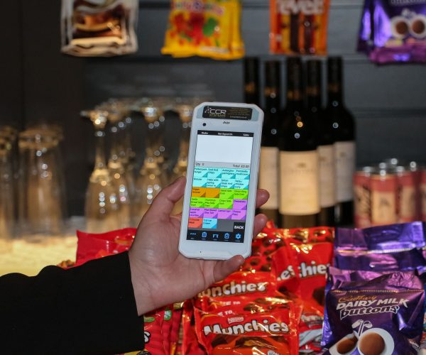 A hand is holding a white device displaying an ordering interface. In the background, there are packets of crisps, bottles of wine, and upside-down glasses on a shelf.