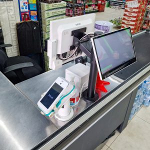 A supermarket checkout counter featuring a till, debit card machine, and a customer display screen. Nearby is a stack of paper bags, a swivel chair, and bottled water on the floor. Shelves stocked with snacks are visible in the background.